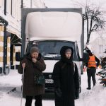 Two women walk on a snowy street near a truck
